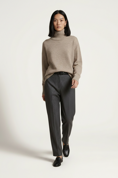 A model stands against a plain light beige studio background wearing a light taupe turtleneck sweater, dark charcoal gray trousers, and black loafers.