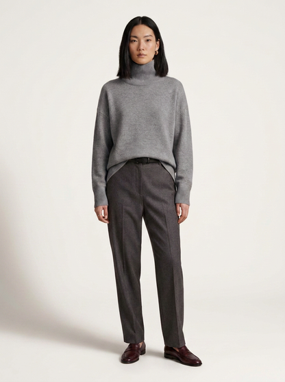 A model stands against a plain light background wearing a light gray turtleneck sweater, dark charcoal gray trousers, and dark brown leather loafers.