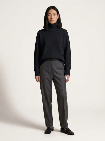 A model stands against a plain light beige studio background wearing a black turtleneck sweater, dark gray trousers, and black loafers.