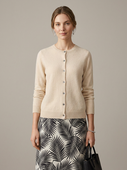 A woman modeling a light beige button-front cardigan sweater paired with a black and white palm leaf print skirt, holding a black handbag against a solid neutral studio background.