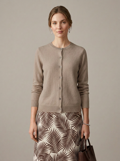 A woman models a taupe knit cardigan sweater with a button-down front, paired with a brown and cream palm-leaf print skirt and holding a dark brown handbag, set against a solid taupe background.