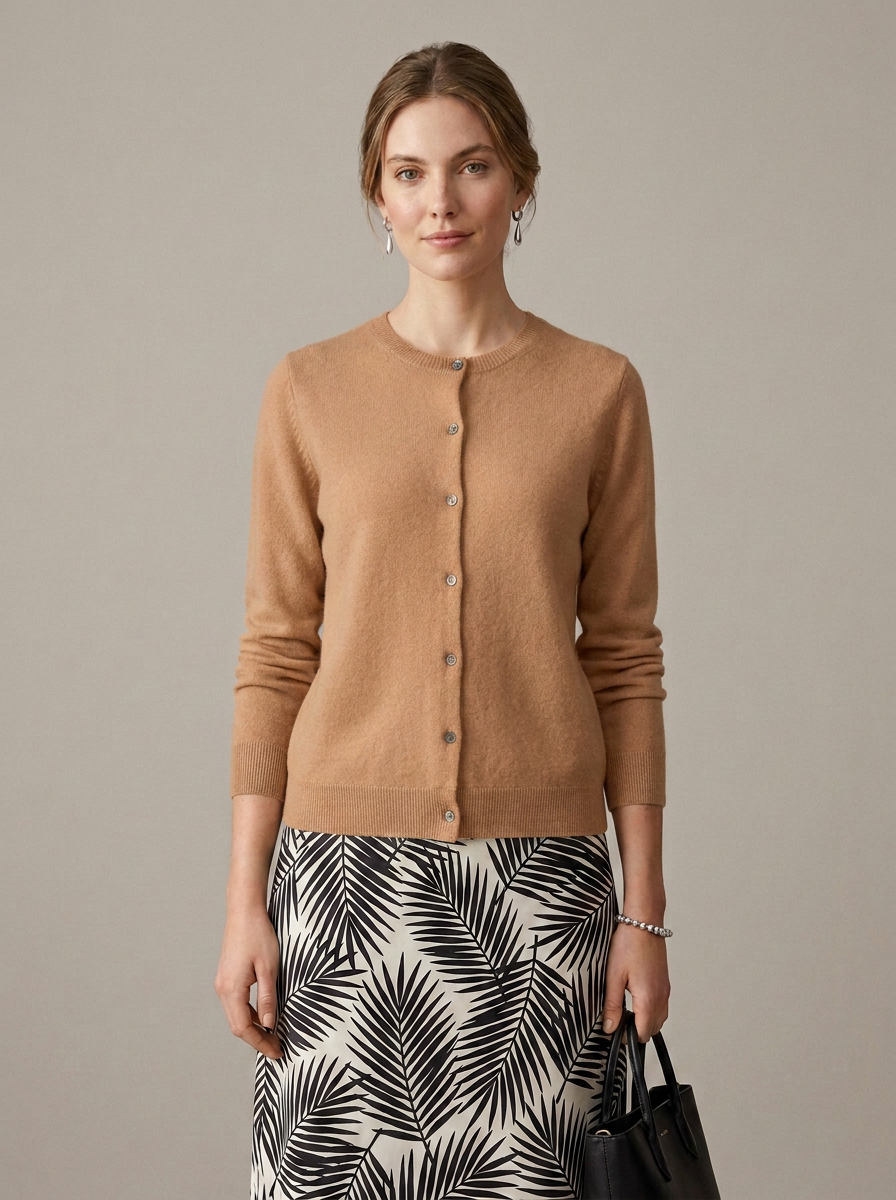 A woman stands against a plain warm gray studio background wearing a tan button-front cardigan over a black-and-white palm leaf print skirt, with a black handbag at her side.