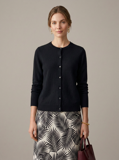 A woman modeling a black long-sleeve button-front cardigan sweater paired with a black and white palm leaf print skirt, holding a burgundy handbag against a solid taupe background.