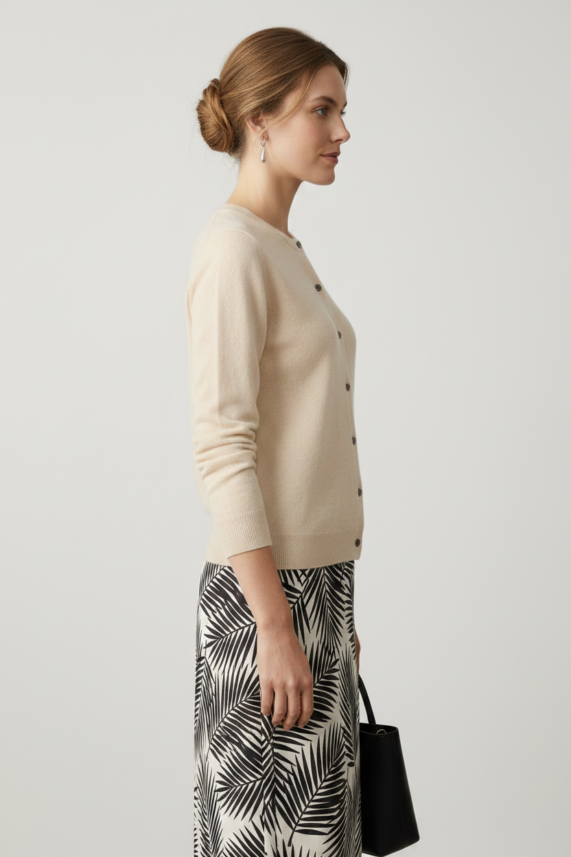 A woman modeling a light beige button-front cardigan sweater paired with a black and white palm leaf print skirt, holding a black handbag against a solid neutral studio background.