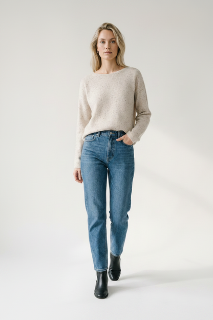A woman stands against a plain light gray studio background wearing a light beige knit sweater, blue jeans, and black ankle boots.