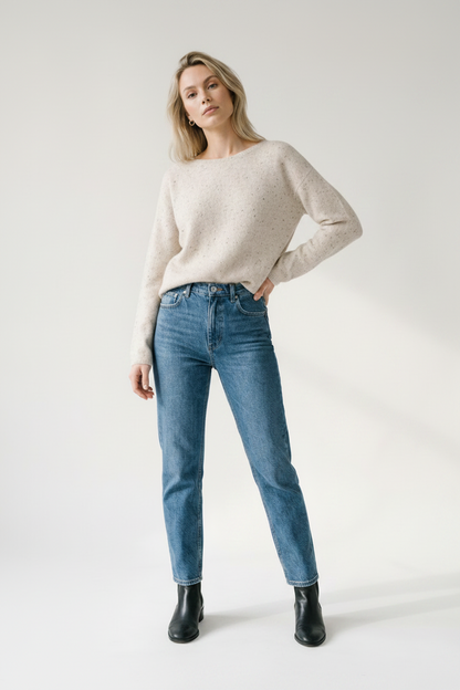 A woman stands against a plain light gray studio background wearing a light beige knit sweater, blue jeans, and black ankle boots.
