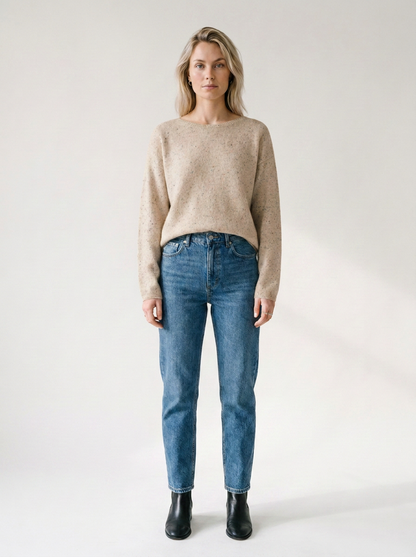 A woman stands against a plain light background wearing a beige knit sweater, blue jeans, and black ankle boots.