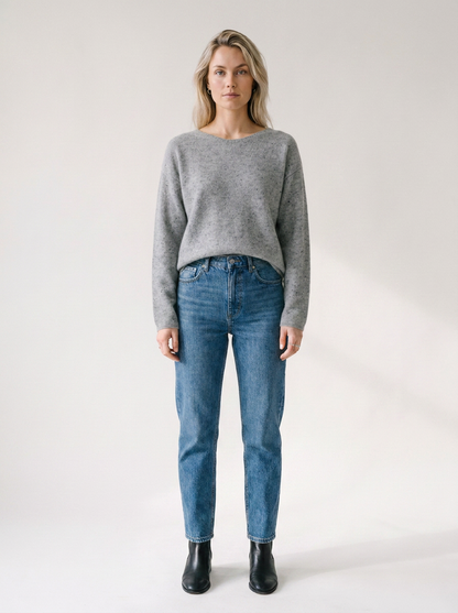 A woman stands against a plain light studio background wearing a heather gray long-sleeve sweater, blue jeans, and black ankle boots.