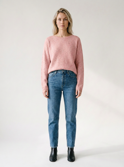 A woman stands against a plain light gray studio background wearing a fuzzy dusty pink long-sleeve sweater, blue jeans, and black ankle boots.