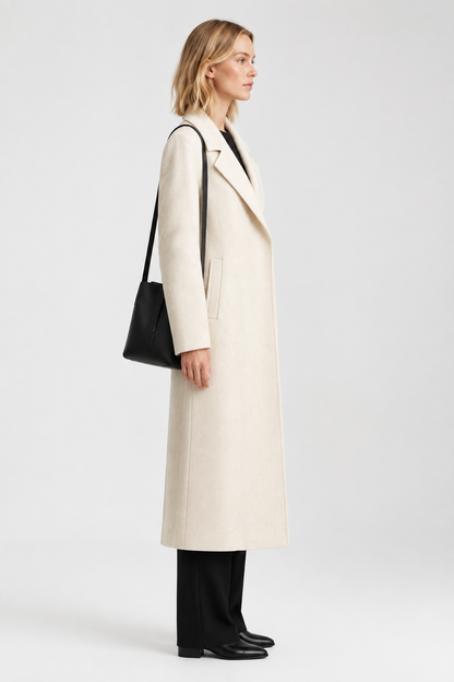 A woman in profile wearing a long cream-colored coat, black trousers, and carrying a black shoulder bag, photographed against a light gray studio background.