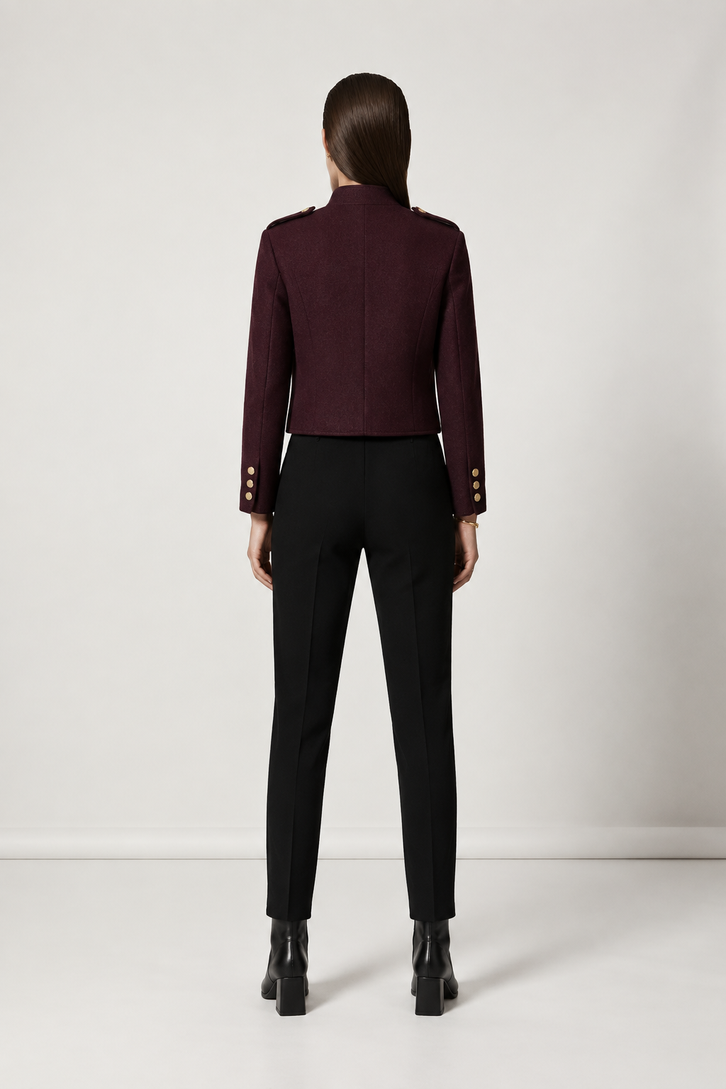 A model shown from the back wearing a cropped, structured burgundy blazer with gold button details on the sleeves, paired with black trousers and black boots, against a plain light gray studio background.