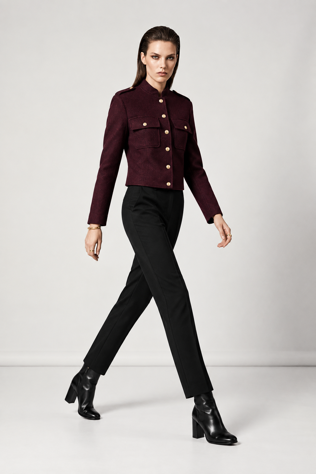 A model walks in a studio wearing a cropped, long-sleeve military-style jacket in deep burgundy with gold buttons, paired with black trousers and black ankle boots.