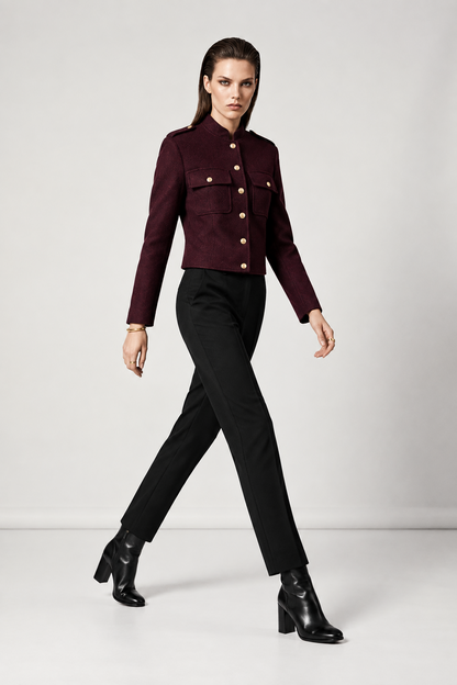 A model walks in a studio wearing a cropped, long-sleeve military-style jacket in deep burgundy with gold buttons, paired with black trousers and black ankle boots.