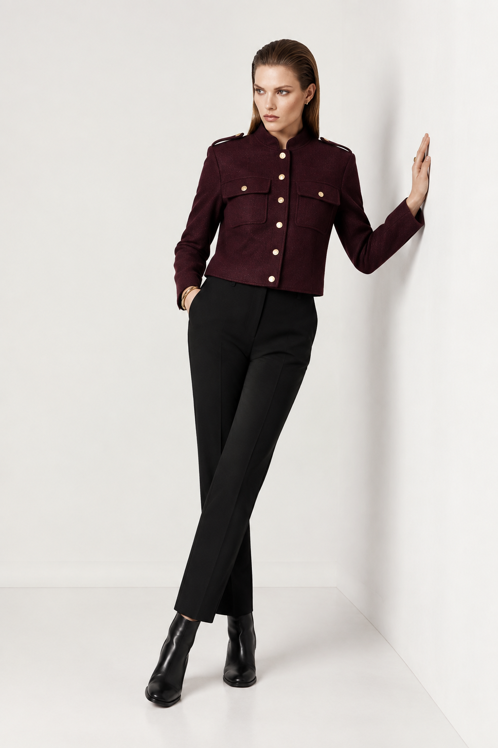 A model stands in a studio corner wearing a cropped dark burgundy military-style jacket with gold buttons, black trousers, and black ankle boots.