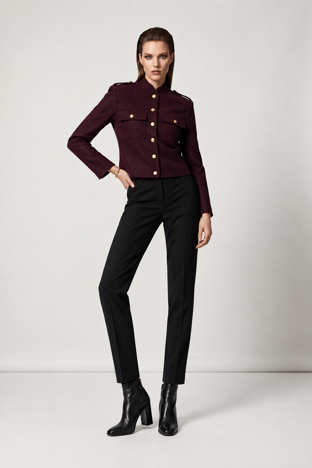 A model stands in a studio wearing a structured, cropped burgundy military-style jacket with gold buttons, paired with black trousers and black heeled ankle boots.