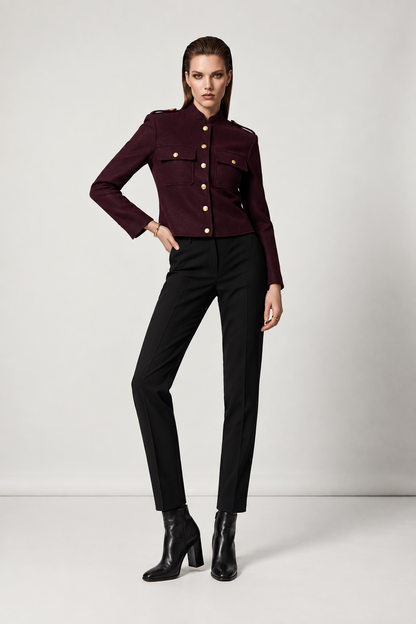 A model stands in a studio wearing a structured, cropped burgundy military-style jacket with gold buttons, paired with black trousers and black heeled ankle boots.