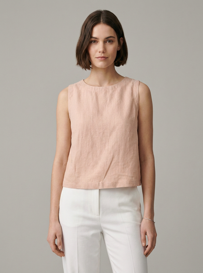 A woman modeling a sleeveless, light blush pink linen top paired with white trousers, shot against a solid gray studio background.