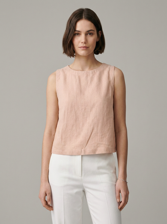 A woman modeling a sleeveless, light blush pink linen top paired with white trousers, shot against a solid gray studio background.