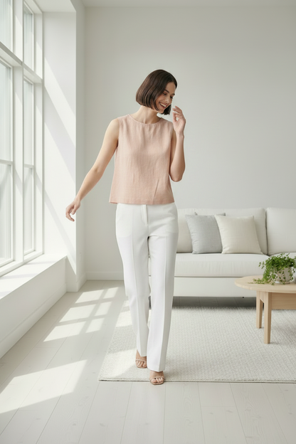 A woman stands in a bright, modern living room wearing a sleeveless blush pink top and white wide-leg trousers with beige high-heeled sandals.
