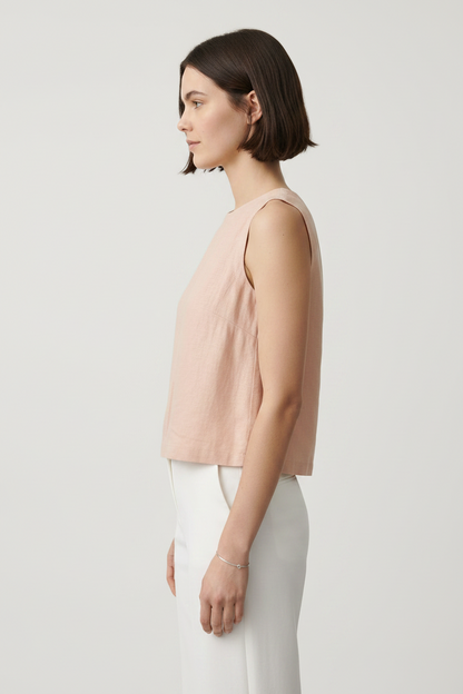 A woman modeling a sleeveless, light blush pink linen top paired with white trousers, shot against a solid gray studio background.
