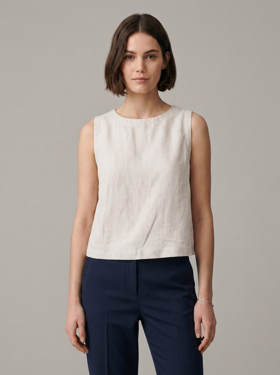 A woman modeling a sleeveless, off-white linen top paired with navy blue trousers, shot against a solid light gray studio background.