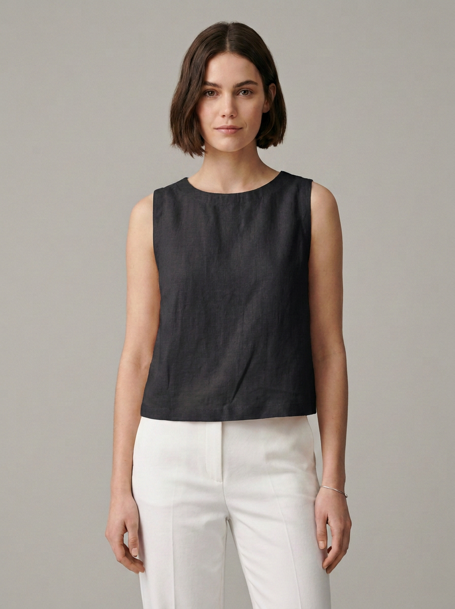A woman modeling a sleeveless black top with a round neckline, paired with white trousers, against a solid gray studio background.