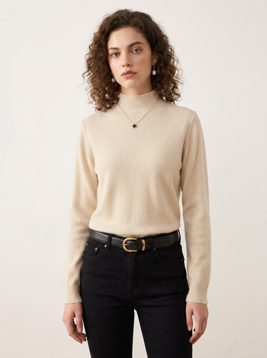 A woman with curly brown hair wearing a beige mock-neck knit sweater, black jeans, and a black belt with a gold buckle, standing against a plain off-white background.