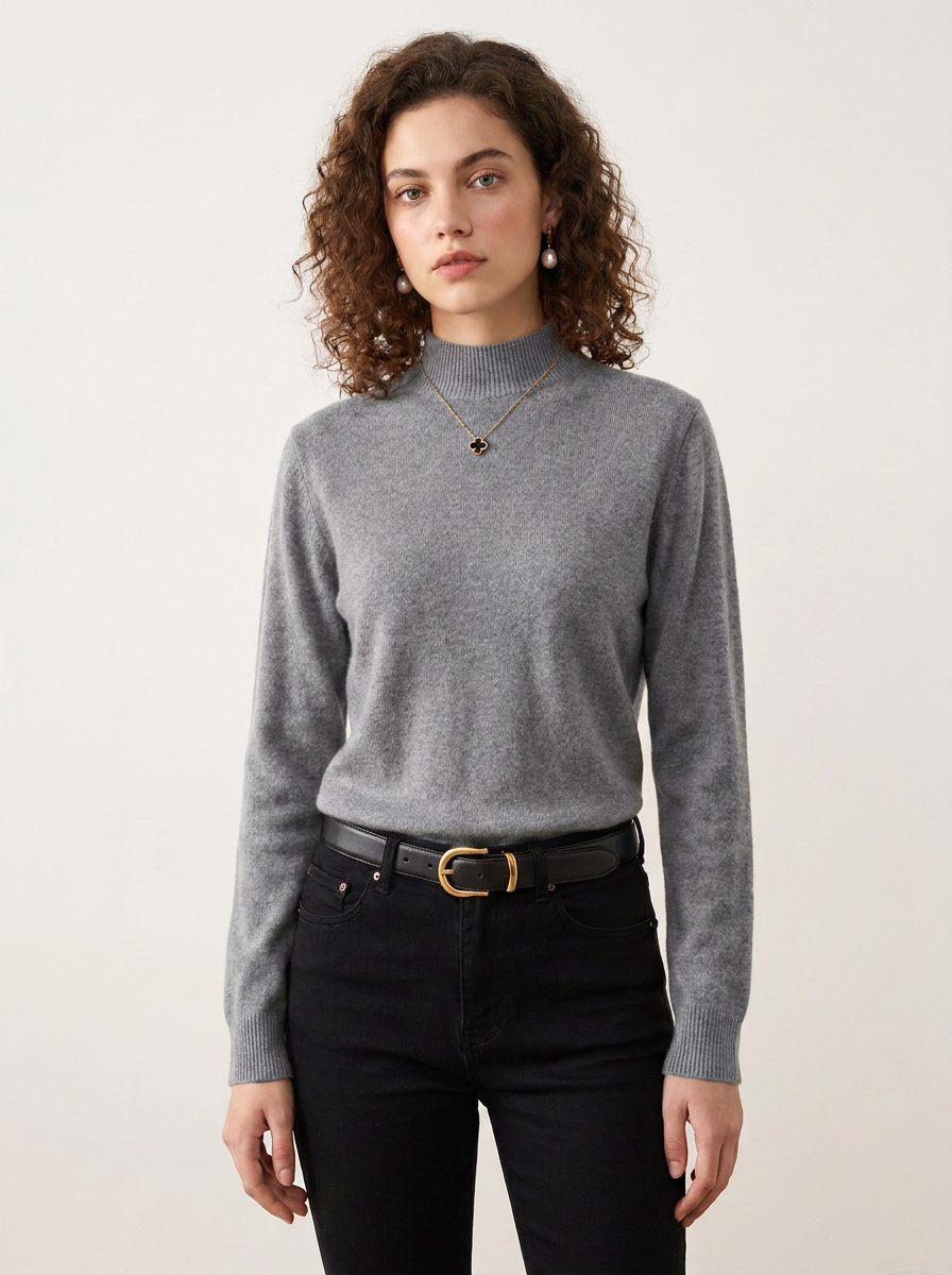A woman with curly brown hair wearing a light gray mock neck knit sweater, black jeans, and a black leather belt with a gold buckle, standing against a plain off-white background.