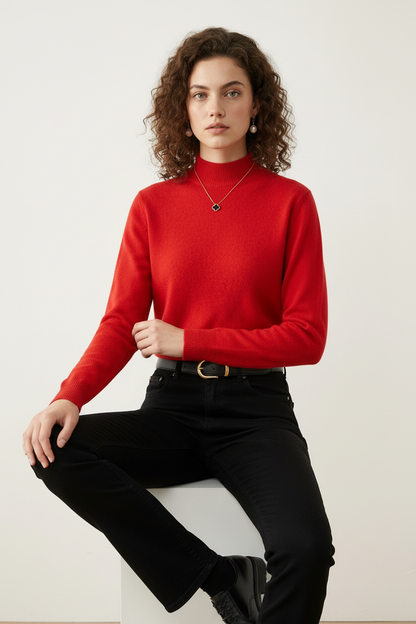 A woman with curly brown hair wearing a bright red mock-neck knit sweater, black jeans, and a black leather belt with a gold buckle, standing against a plain off-white background.