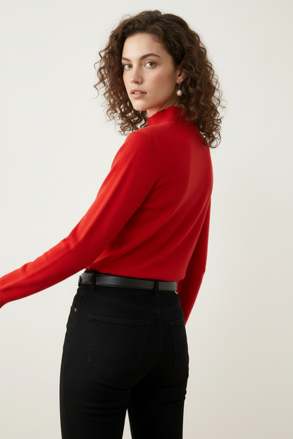 A woman with curly brown hair wearing a bright red mock-neck knit sweater, black jeans, and a black leather belt with a gold buckle, standing against a plain off-white background.