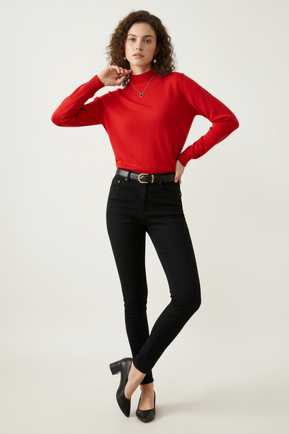 A woman with curly brown hair wearing a bright red mock-neck knit sweater, black jeans, and a black leather belt with a gold buckle, standing against a plain off-white background.