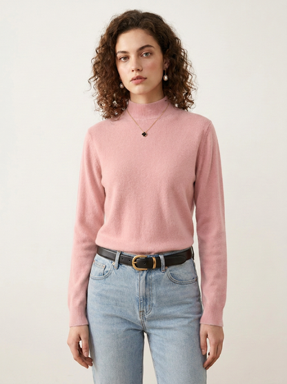 A woman with curly brown hair wearing a dusty pink mock neck knit sweater, light wash blue jeans, and a black leather belt with a gold buckle, standing against a plain off-white background.