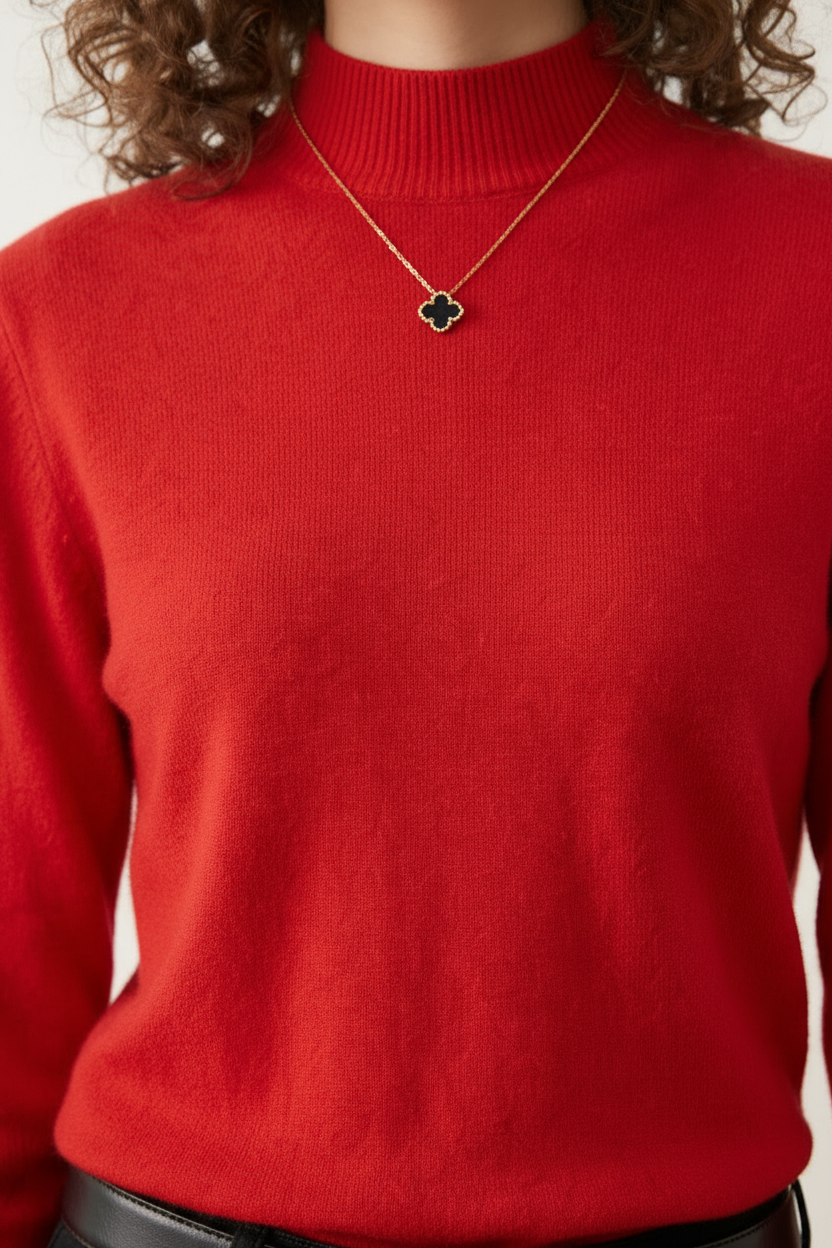A woman with curly brown hair wearing a bright red mock-neck knit sweater, black jeans, and a black leather belt with a gold buckle, standing against a plain off-white background.