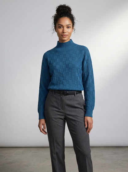 A woman modeling a blue textured mock-neck sweater paired with dark gray trousers, standing against a neutral light gray studio background.