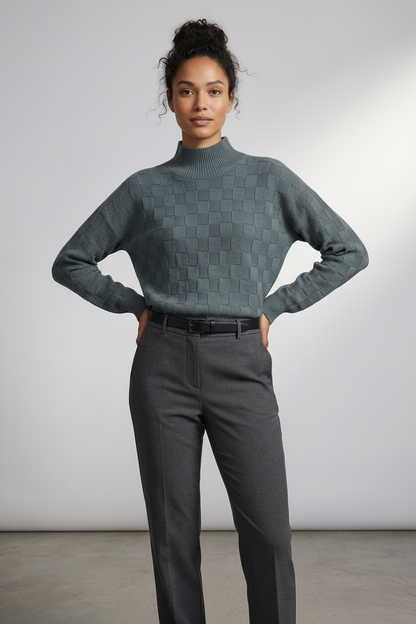 A woman modeling a slate blue-green textured mock-neck sweater paired with dark gray trousers, standing against a neutral light gray studio background.