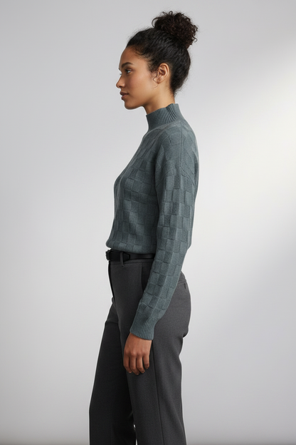 A woman modeling a slate blue-green textured mock-neck sweater paired with dark gray trousers, standing against a neutral light gray studio background.
