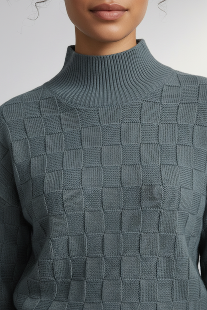 A woman modeling a slate blue-green textured mock-neck sweater paired with dark gray trousers, standing against a neutral light gray studio background.