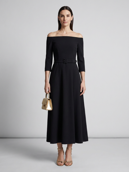 A woman models an off-the-shoulder black midi dress with three-quarter sleeves and a matching belt. She is holding a small metallic gold handbag and wearing gold strappy heels, set against a neutral light gray studio background.