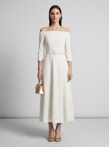 A woman models an off-the-shoulder midi dress in off-white with a matching belt and silver buckle. She is holding a small metallic gold handbag and wearing gold strappy heels, set against a neutral light gray studio background.