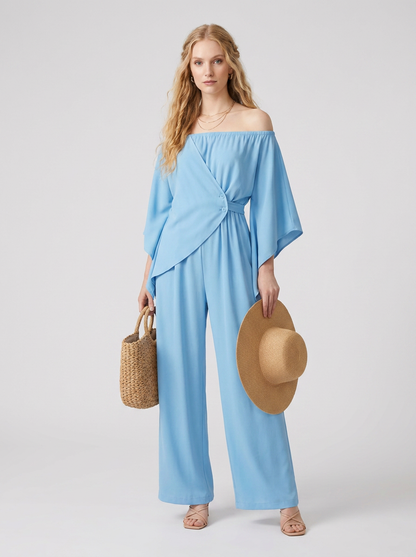 A woman modeling a light blue off-the-shoulder wrap-style jumpsuit with wide legs, holding a woven straw hat and a small woven tote bag against a plain light gray studio background.