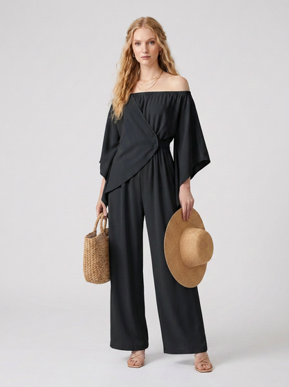 A woman modeling an off-the-shoulder black wide-leg jumpsuit, holding a straw hat and a woven straw tote bag, against a light gray studio background.