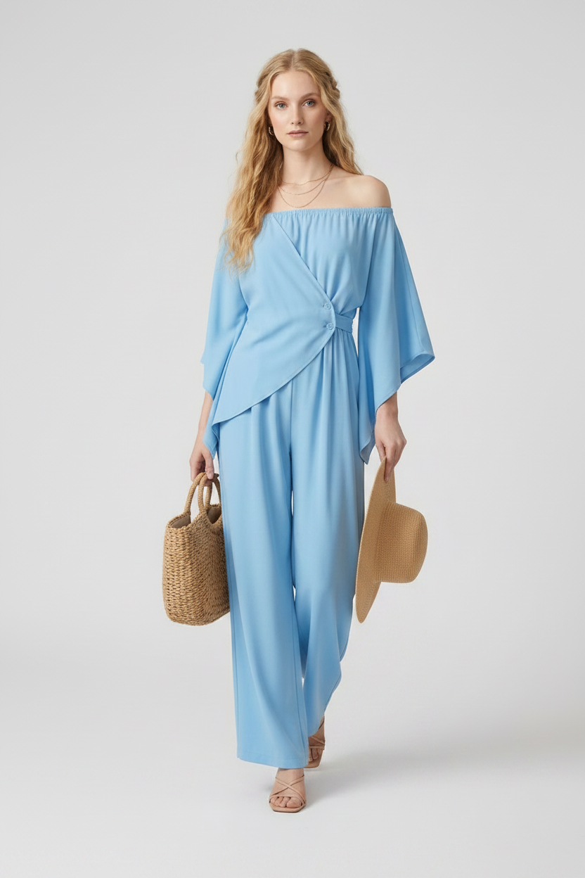 A woman modeling a light blue off-the-shoulder wrap-style jumpsuit with wide legs, holding a woven straw hat and a small woven tote bag against a plain light gray studio background.