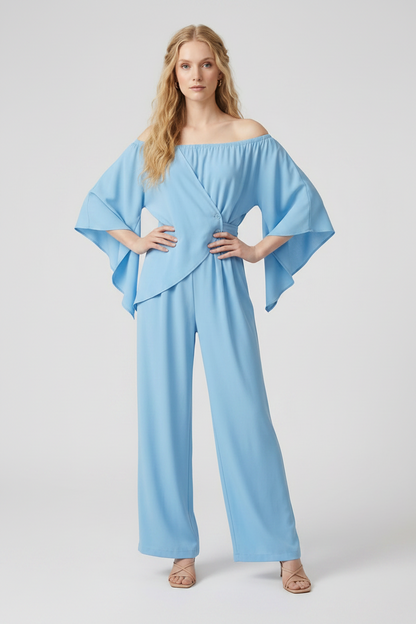 A woman modeling a light blue off-the-shoulder wrap-style jumpsuit with wide legs, holding a woven straw hat and a small woven tote bag against a plain light gray studio background.