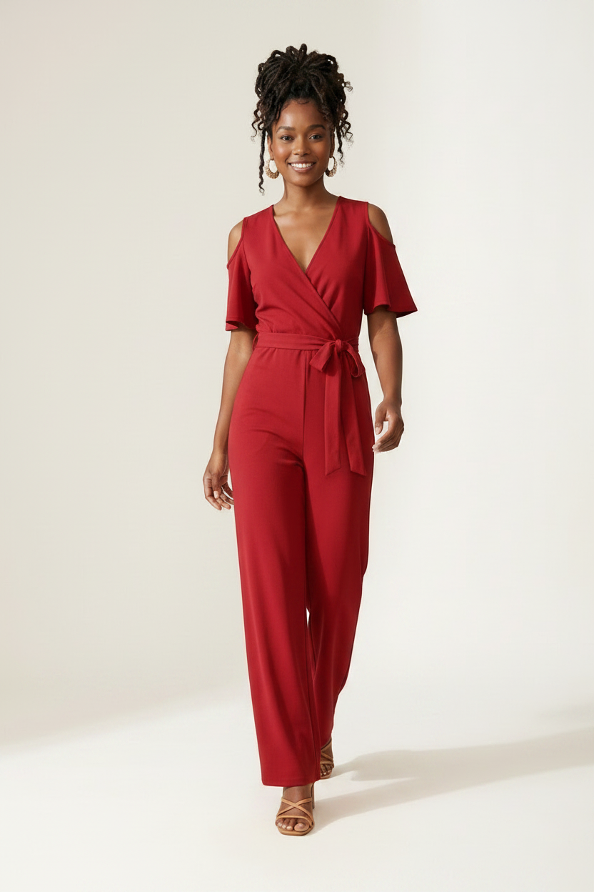 A woman modeling a red cold-shoulder wrap jumpsuit with wide legs and a tie waist, holding a woven straw tote bag against a plain off-white studio background.