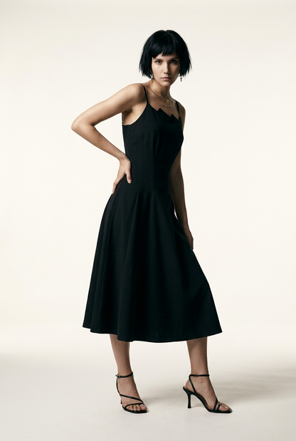 A model with a short dark bob hairstyle wearing a black midi-length spaghetti strap dress with a unique scalloped neckline, paired with black strappy high-heeled sandals, standing against a warm off-white studio background.