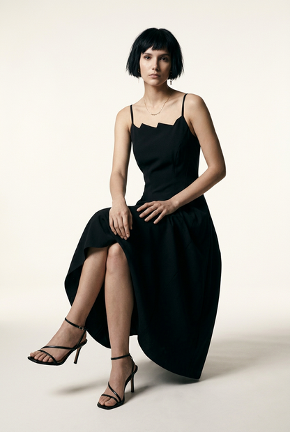 A model with a short dark bob hairstyle wearing a black midi-length spaghetti strap dress with a unique scalloped neckline, paired with black strappy high-heeled sandals, standing against a warm off-white studio background.