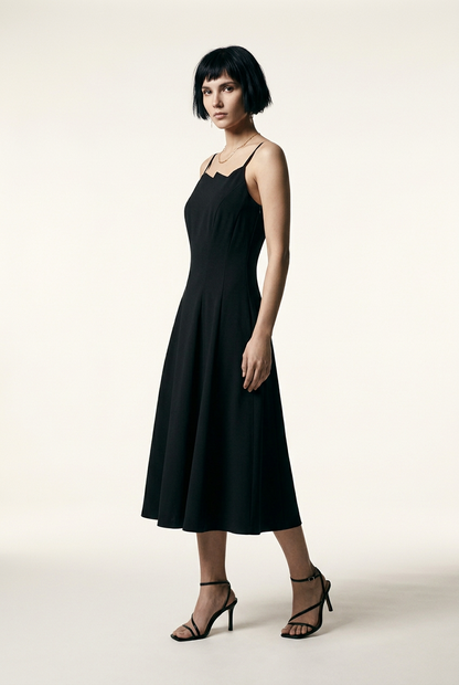 A model with a short dark bob hairstyle wearing a black midi-length spaghetti strap dress with a unique scalloped neckline, paired with black strappy high-heeled sandals, standing against a warm off-white studio background.