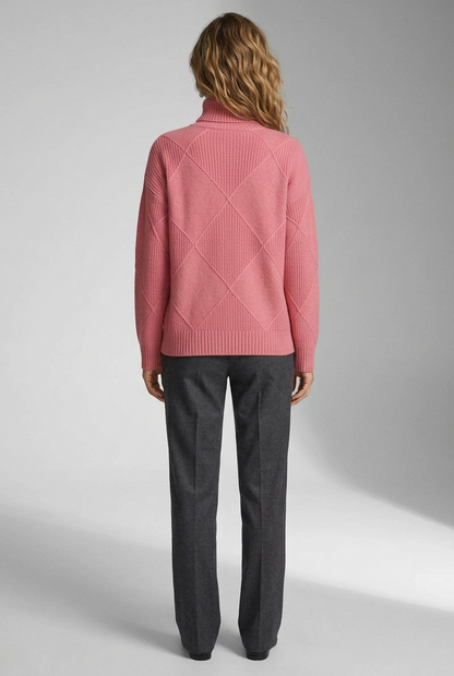 A woman modeling a pink ribbed turtleneck sweater with a diamond-shaped argyle knit pattern, paired with dark gray trousers, against a neutral gray studio background.