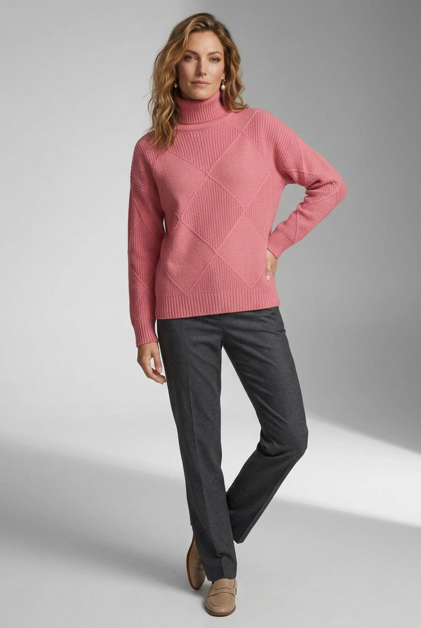 A woman modeling a pink ribbed turtleneck sweater with a diamond-shaped argyle knit pattern, paired with dark gray trousers, against a neutral gray studio background.