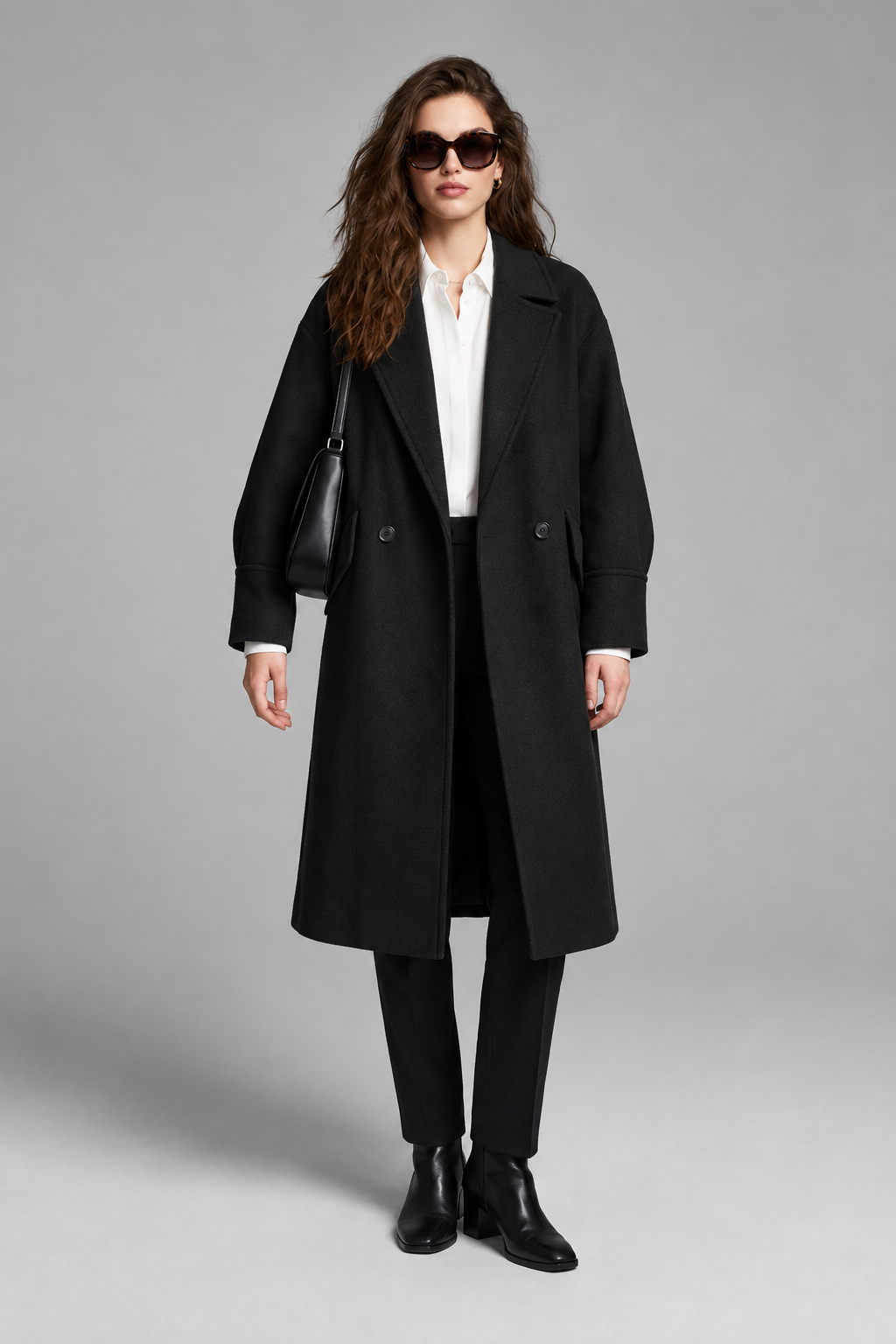A woman models a long camel-colored double-breasted coat over a white collared shirt, black trousers, and black ankle boots, while wearing black sunglasses and carrying a black shoulder bag against a neutral gray studio background.